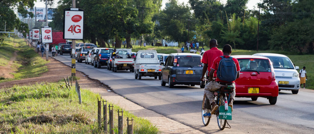Two boys on a bicycle and car traffic passing several Airtel advertisements in Malawi